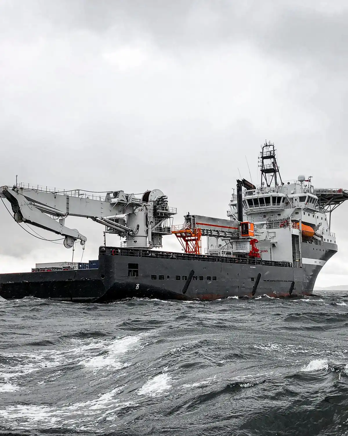 A grey working vessel on open sea under a pale sky, deck equipment visible along its side.