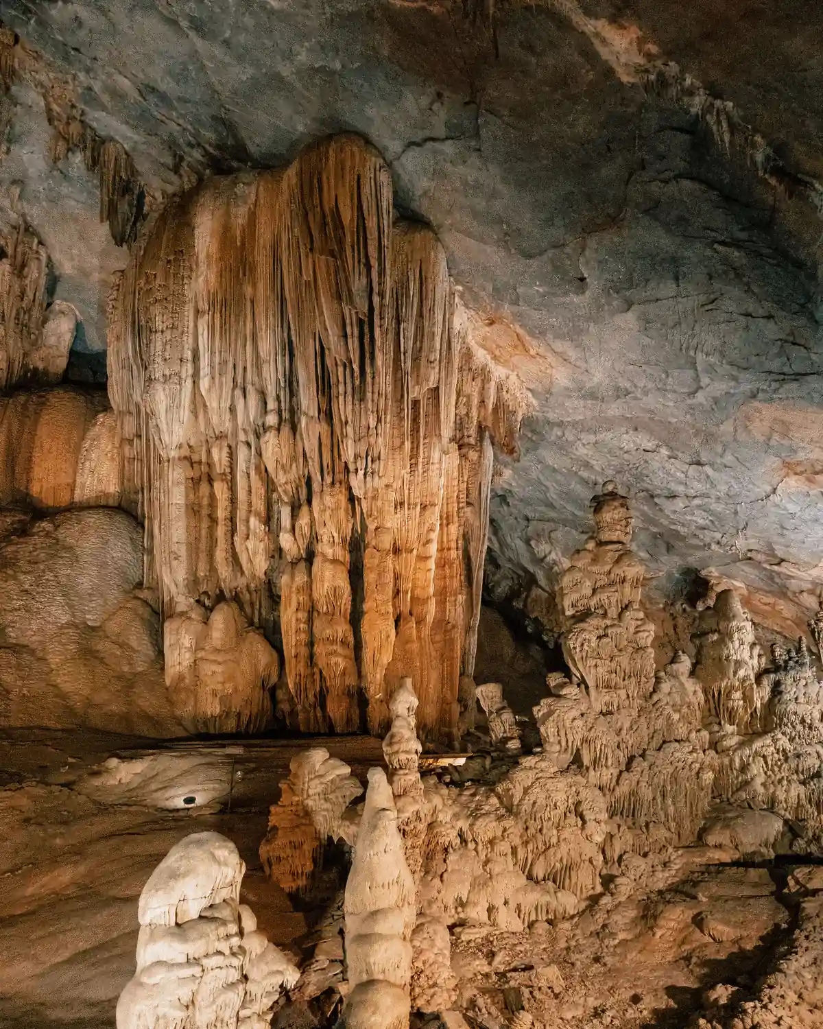 Pale stalactites and flowstone hanging from a cave roof above rough rock on the cave floor.