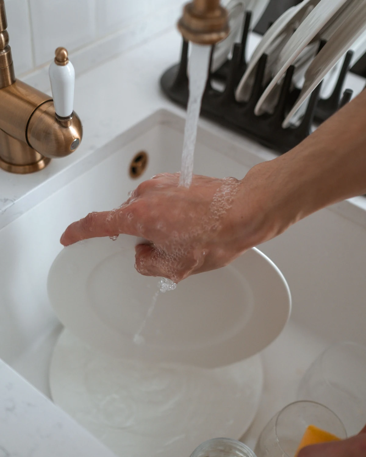 Hands washing a white ceramic plate under running water at a kitchen sink.