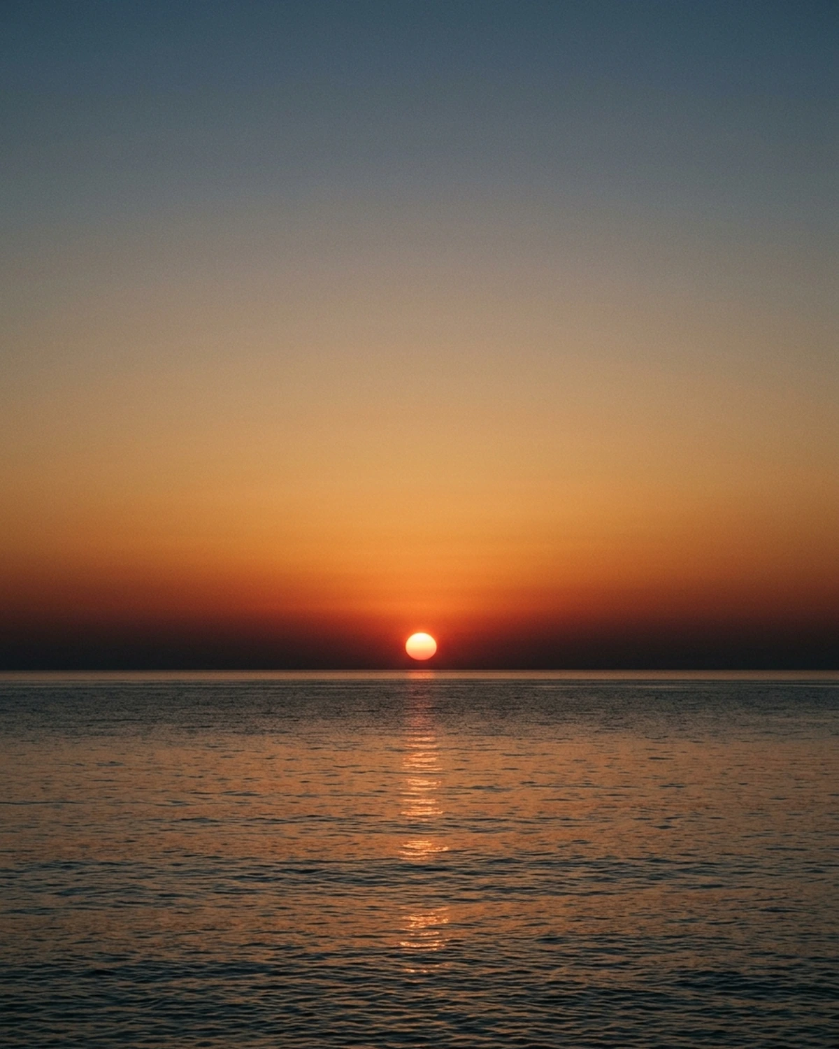 A red-orange Sun setting over a calm ocean beneath a sky fading to blue